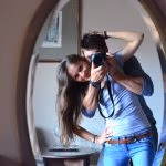A smilling girl looking in to a mirror with a man huging her from behind holding a camera pointing at the mirror.