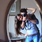 A smilling girl looking in to a mirror with a man huging her from behind holding a camera pointing at the mirror.