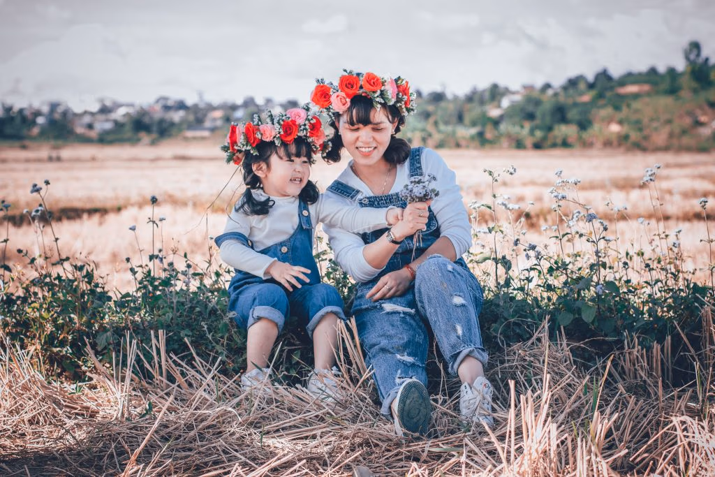 Adult women waring jeans and flower in her hair sitting in the gras next to a identical dressed child. Both are smiling happily in the shades on a sunny day outside.