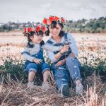 Adult women waring jeans and flower in her hair sitting in the gras next to a identical dressed child. Both are smiling happily in the shades on a sunny day outside.