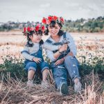 Adult women waring jeans and flower in her hair sitting in the gras next to a identical dressed child. Both are smiling happily in the shades on a sunny day outside.