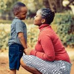 African women in a white and black skirt with a red shirt, kneeling infront of a child in blue shorts and a green t-shirt, both are smiling lovingly