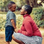 African women in a white and black skirt with a red shirt, kneeling infront of a child in blue shorts and a green t-shirt, both are smiling lovingly