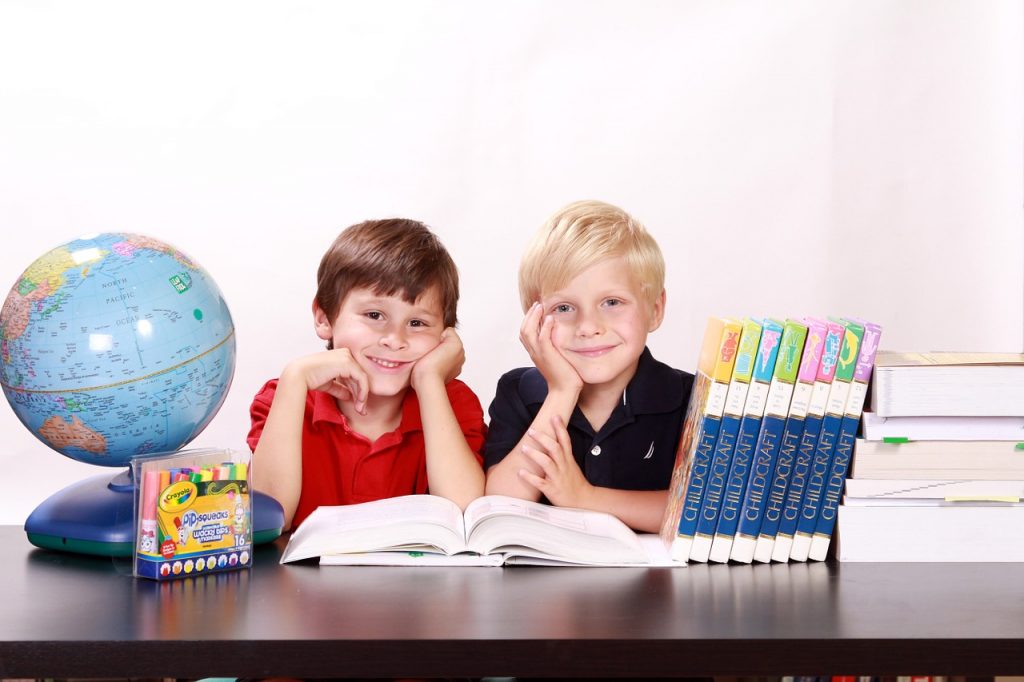 Two boys sitting next to each other in front of a desk with books and a globe on the table